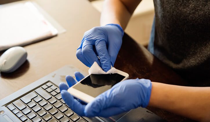 Man with gloves hands wiping mobile phone with disinfectant wipe while working from home. Horizontal indoors close-up with copy space.