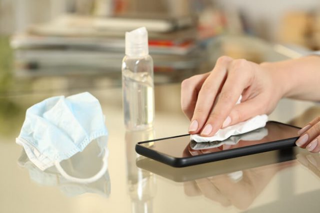 Close up of girl hand with protective mask disinfecting with sanitizer gel her phone screen preventing contagion sitting on a desk at home