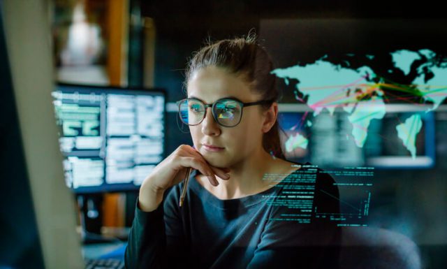 Stock image of a young woman, wearing glasses, surrounded by computer monitors in a dark office. In front of her there is a see-through displaying telecom technology trends