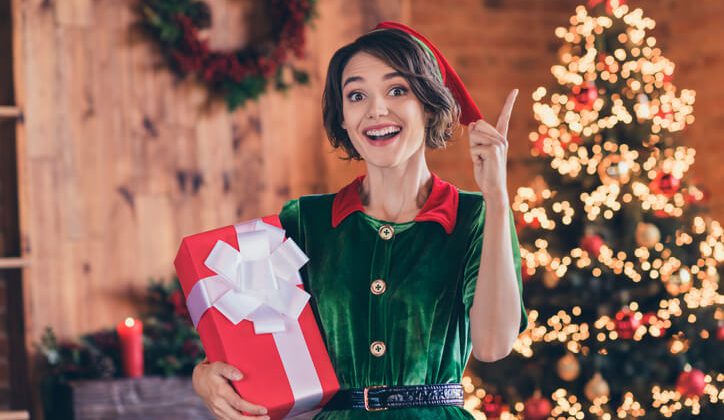 Photo of sweet smart young lady wear elf clothes holding present celebrating christmas pointing finger smiling indoors house home room