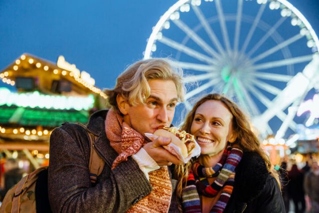 Happy couple are at the christmas fair and market. The man is eating a hotdog from one of the market stalls and his partner is laughing.