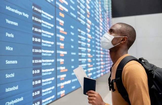 Portrait of a black male traveler wearing a facemask at the airport and looking at the flight schedule - travel essentials