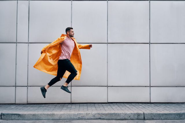 Trendy young man in yellow raincoat jumping in the air on wall street urban background. carefree, freedom, escape concept
