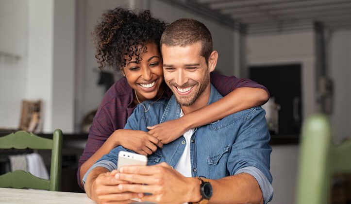 Smiling young couple embracing while looking at smartphone. Multiethnic couple sharing social media on smart phones while sitting at table. Smiling african girl embracing from behind her happy boyfriend while using cellphone and laughing.
