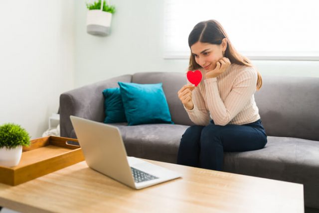 Good-looking woman in love is in the living room sending love messages and showing a red heart to his boyfriend or partner during a video call in her laptop using Long-Distance Relationship 