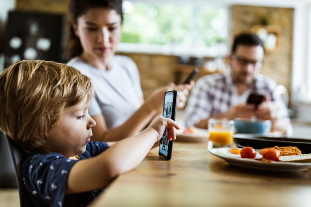 Profile view of small boy using smart phone at dining table while his parents are in the background. parental control idea image