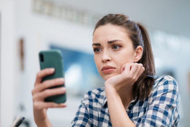 Close up of a depressed Caucasian young woman sitting at home, reading some bad news on her smart phone and health