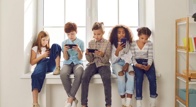 Group of children of different nationalities sit in a row on the windowsill and with lack of emotion play online games or read social networks on mobile phones