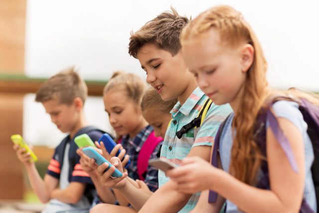 group of happy elementary school students with smartphones and backpacks sitting on bench outdoors